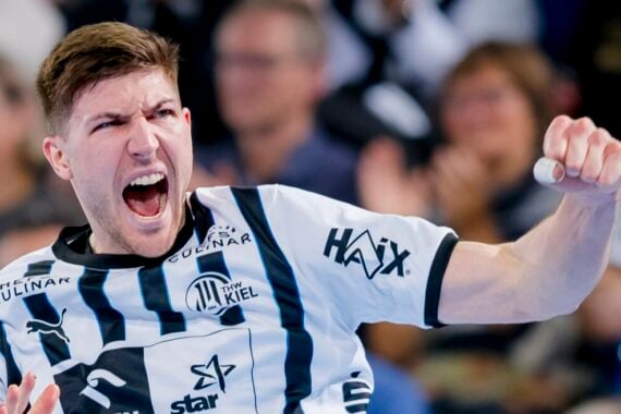 Ein männlicher Handballspieler in einem schwarz-weiß gestreiften Trikot jubelt energisch auf dem Spielfeld, während im Hintergrund eine begeisterte Menge steht.