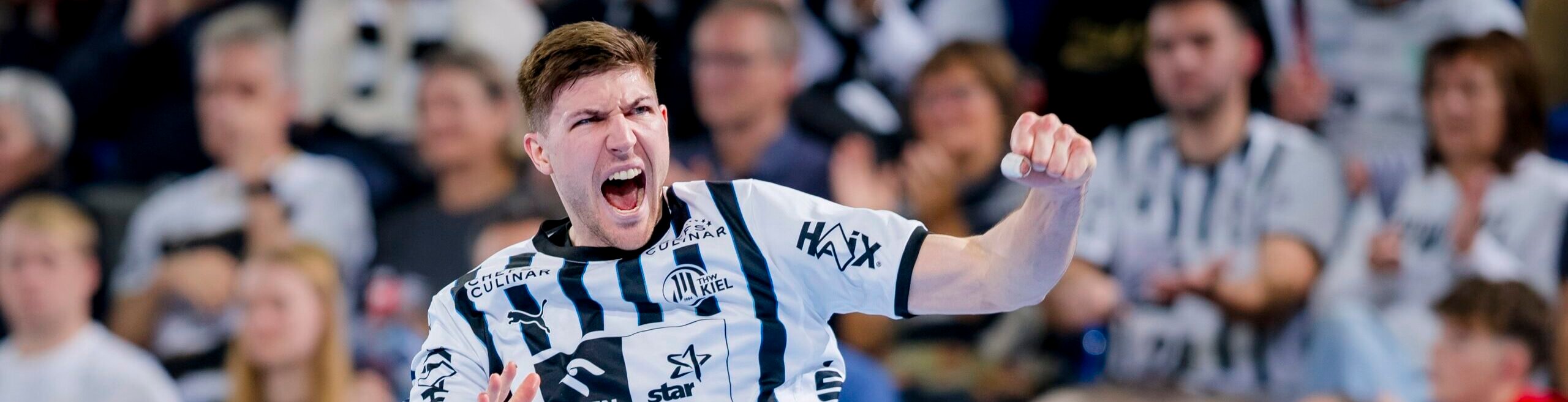 Ein männlicher Handballspieler in einem schwarz-weiß gestreiften Trikot jubelt energisch auf dem Spielfeld, während im Hintergrund eine begeisterte Menge steht.