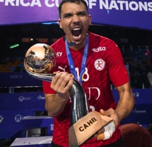 Ein männlicher Sportler in einem roten Trikot jubelt, während er eine große Trophäe mit der Aufschrift CAHB in der Hand hält, in einer Halle mit einem blauen Banner des Africa Cup of Nations im Hintergrund.