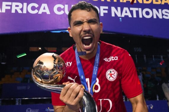 Ein männlicher Athlet in einem roten Trikot feiert ausgelassen, hält eine goldene Trophäe mit einem gebogenen Griff und trägt eine blaue Siegermedaille. Hinter ihm prangt ein Banner mit der Aufschrift AFRICA CUP OF NATIONS.