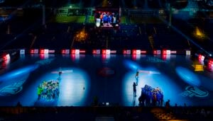 Eine hell erleuchtete Sporthalle mit zwei Gruppen von Menschen auf dem Spielfeld, die von Scheinwerfern angestrahlt werden, und einer Anzeigetafel über den leeren Zuschauerplätzen.