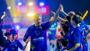 Eine Gruppe von Sportlern in blauen Uniformen klatscht sich auf einem hell erleuchteten Hallenplatz gegenseitig ab und feiert gemeinsam. Mehrere Kinder sind ebenfalls anwesend und nehmen an der Feier teil.