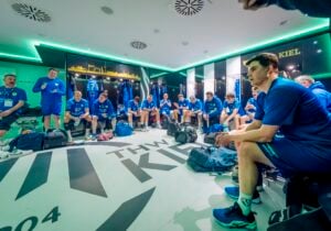 Eine Herren-Handballmannschaft in blauen Trikots sitzt in einer modernen Umkleidekabine und hört einem Trainer zu. Sporttaschen liegen auf dem Boden verstreut, und auf dem Boden steht THW KIEL geschrieben. Die Atmosphäre ist konzentriert und ernst.