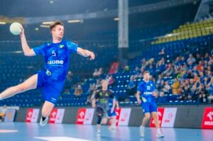 Ein männlicher Handballspieler in einem blauen Trikot springt in die Luft und bereitet sich darauf vor, den Ball während eines Spiels in einer Halle zu werfen, während im Hintergrund Mitspieler, Gegner und Zuschauer zu sehen sind.