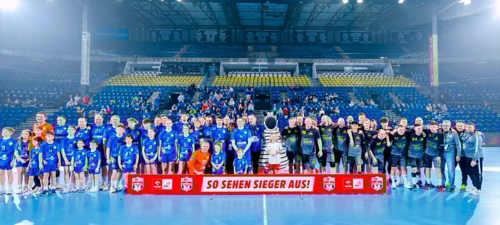 HANDBALL 2025/26: Aktion „Euer Topspiel“ Eine große Gruppe von Handballspielern, Trainern, Kindern und einem Maskottchen posieren zusammen auf einem Hallenplatz, mit leeren gelben und blauen Stadionsitzen und Zuschauern im Hintergrund. Auf einem Banner davor steht SO SEHEN SIEGER AUS!.