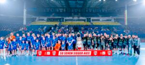 Eine große Gruppe von Handballspielern, Trainern, Kindern und einem Maskottchen posieren zusammen auf einem Hallenplatz, mit leeren gelben und blauen Stadionsitzen und Zuschauern im Hintergrund. Auf einem Banner davor steht SO SEHEN SIEGER AUS!.