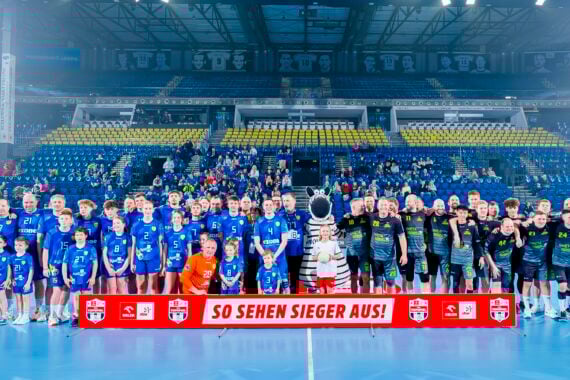 Eine große Gruppe von Handballspielern, Trainern, Kindern und einem Maskottchen posieren zusammen auf einem Hallenplatz, mit leeren gelben und blauen Stadionsitzen und Zuschauern im Hintergrund. Auf einem Banner davor steht SO SEHEN SIEGER AUS!.