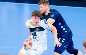 Zwei männliche Handballspieler in Aktion auf dem Spielfeld während eines spannenden Derbys zwischen dem THW Kiel und der SG Flensburg-Handewitt; der Spieler in Weiß hält den Ball und versucht, an dem Abwehrspieler in Blau vorbeizukommen, der ihm den Weg versperrt.
