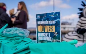 Auf einem Tisch mit grünen Handschuhen liegt ein Werbeflyer für einen Müllsammelwettbewerb aus. Menschen und eine Person in einem gestreiften Kostüm sind im Hintergrund unter einem blauen Himmel unscharf zu erkennen.