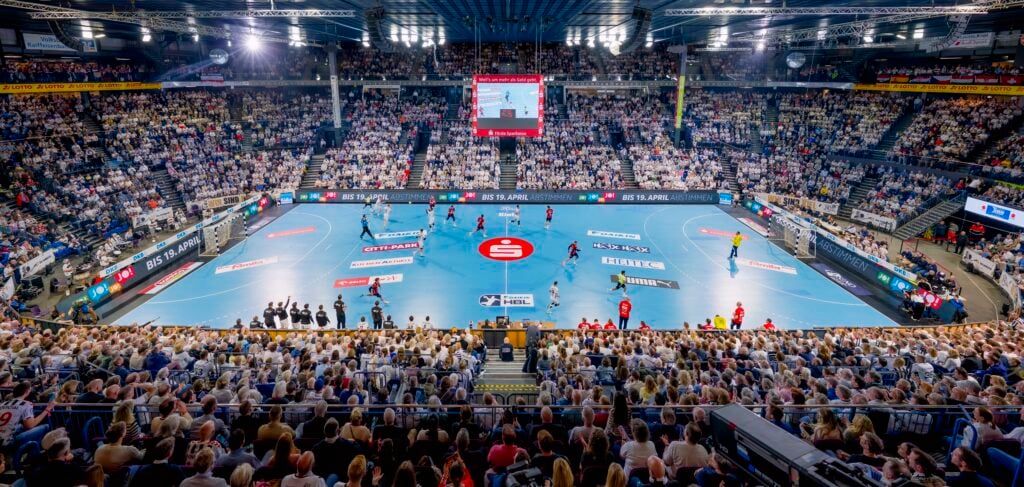 HANDBALL 2025/26: THW Kiel - SG Flensburg-Handewitt Panoramablick auf ein überfülltes Hallenstadion während eines Handballspiels, mit Spielern auf einem blauen Spielfeld und Zuschauern auf den Tribünen. Große Bildschirme und helle Lichter erhellen die lebhafte Szene.