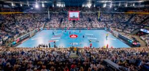Panoramablick auf ein überfülltes Hallenstadion während eines Handballspiels, mit Spielern auf einem blauen Spielfeld und Zuschauern auf den Tribünen. Große Bildschirme und helle Lichter erhellen die lebhafte Szene.