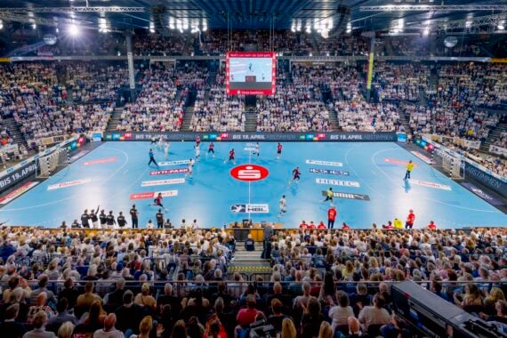 Panoramablick auf ein überfülltes Hallenstadion während eines Handballspiels, mit Spielern auf einem blauen Spielfeld und Zuschauern auf den Tribünen. Große Bildschirme und helle Lichter erhellen die lebhafte Szene.