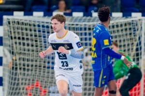 Ein männlicher Handballspieler in einem weißen Trikot mit der Nummer 23 läuft vor dem Tor, während ein anderer Spieler in einem blauen Trikot und ein Torwart im Hintergrund auf einem Hallenplatz stehen.