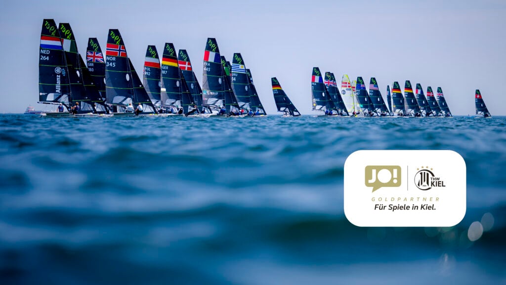 Eine Flotte von Segelbooten mit verschiedenen Nationalflaggen an den Segeln fährt auf dem Meer unter klarem Himmel. Im Vordergrund ist das Logo von JO! und Kiel zu sehen, die als Goldpartner für das Kieler Segelevent gekennzeichnet sind.