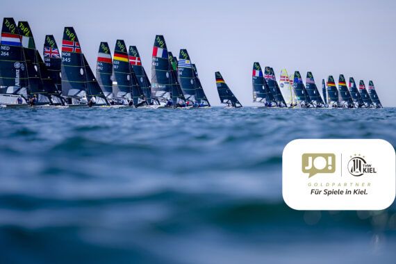 Eine Flotte von Segelbooten mit verschiedenen Nationalflaggen an den Segeln fährt auf dem Meer unter klarem Himmel. Im Vordergrund ist das Logo von JO! und Kiel zu sehen, die als Goldpartner für das Kieler Segelevent gekennzeichnet sind.