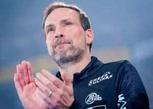 Ein Mann mit kurzen Haaren und gestutztem Bart steht in einem Innenraum, trägt eine schwarze Sportjacke mit weißen Logos und klatscht, während er mit nachdenklicher Miene leicht nach oben schaut.