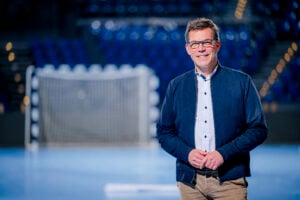 Ein Mann mit Brille, blauer Jacke und khakifarbener Hose steht lächelnd in einer Sporthalle mit einem verschwommenen Handballtor und blauen Sitzgelegenheiten im Hintergrund.