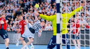 Ein Handballspieler in einem weiß-schwarzen Trikot springt, um den Ball in Richtung Tor zu werfen, während ein Torwart in einem gelben Trikot versucht, den Schuss zu blocken. Andere Spieler und die Zuschauer im Hintergrund schauen aufmerksam zu.