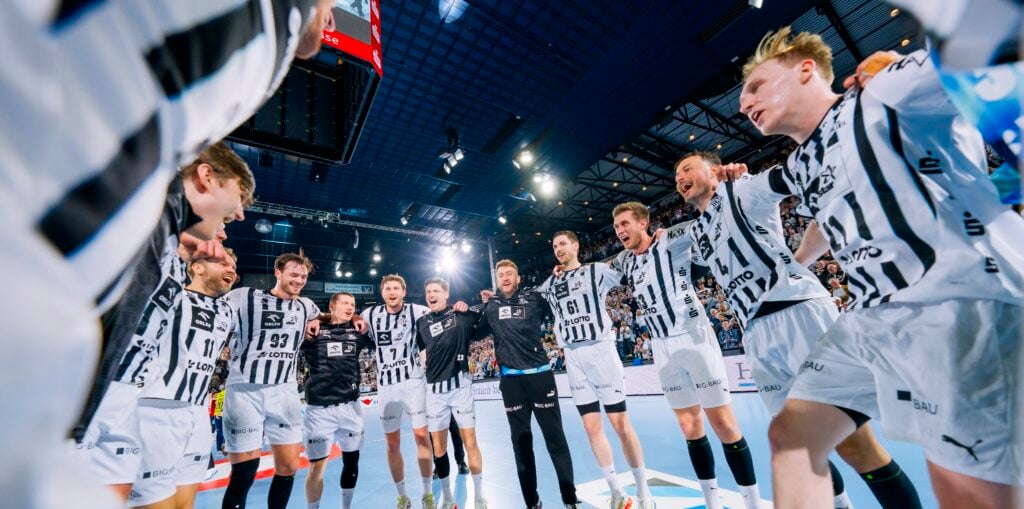 HANDBALL 2025/26: THW Kiel - SG Flensburg-Handewitt Eine Gruppe männlicher Handballspieler in schwarz-weißen Trikots steht mit verschränkten Armen auf einem Hallenplatz im Scheinwerferlicht und feiert.