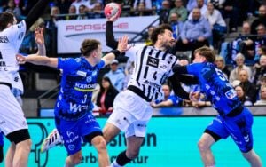 Drei männliche Handballspieler liefern sich während eines Spiels einen intensiven Wettkampf. Ein Spieler in einem weiß gestreiften Trikot springt und hält den Ball, während zwei Spieler in blauen Trikots ihn verteidigen. Die Zuschauer schauen von den Tribünen im Hintergrund zu.