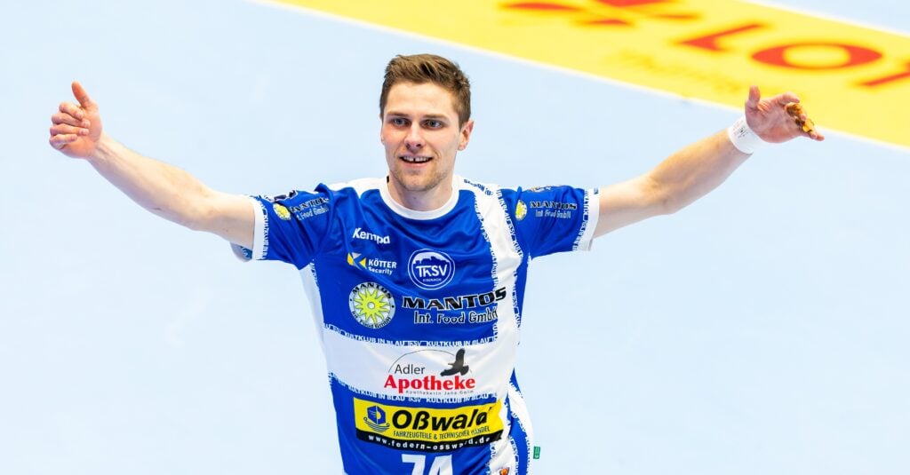 Ein männlicher Handballspieler, der ein blau-weißes Trikot mit Sponsorenlogos trägt, jubelt mit erhobenen Armen auf einem Hallenplatz.