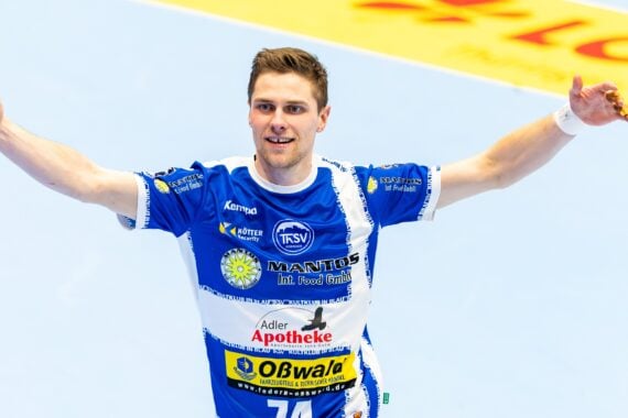 Ein männlicher Handballspieler, der ein blau-weißes Trikot mit Sponsorenlogos trägt, jubelt mit erhobenen Armen auf einem Hallenplatz.