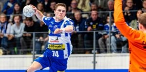 Ein Handballspieler in einem blau-weißen Trikot bereitet sich darauf vor, den Ball während eines Spiels zu werfen, während ein Torwart in einem orangefarbenen Trikot einen Arm zum Abwehren hebt. Die Zuschauer schauen von der Tribüne im Hintergrund zu.