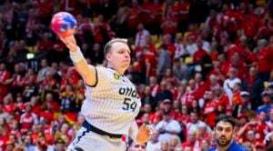 Ein Handballspieler in einem weiß gestreiften Trikot springt und bereitet sich darauf vor, den Ball während eines Spiels zu werfen, während eine rot gekleidete Menge von der Tribüne aus zusieht. Ein anderer Spieler in Blau ist im Vordergrund zu sehen.