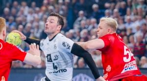 Ein Handballspieler in einem weißen Trikot versucht, den Ball zu werfen, während er von einem Spieler in einem roten Trikot während eines rasanten Spiels eng verteidigt wird, mit einer unscharfen Menschenmenge im Hintergrund.