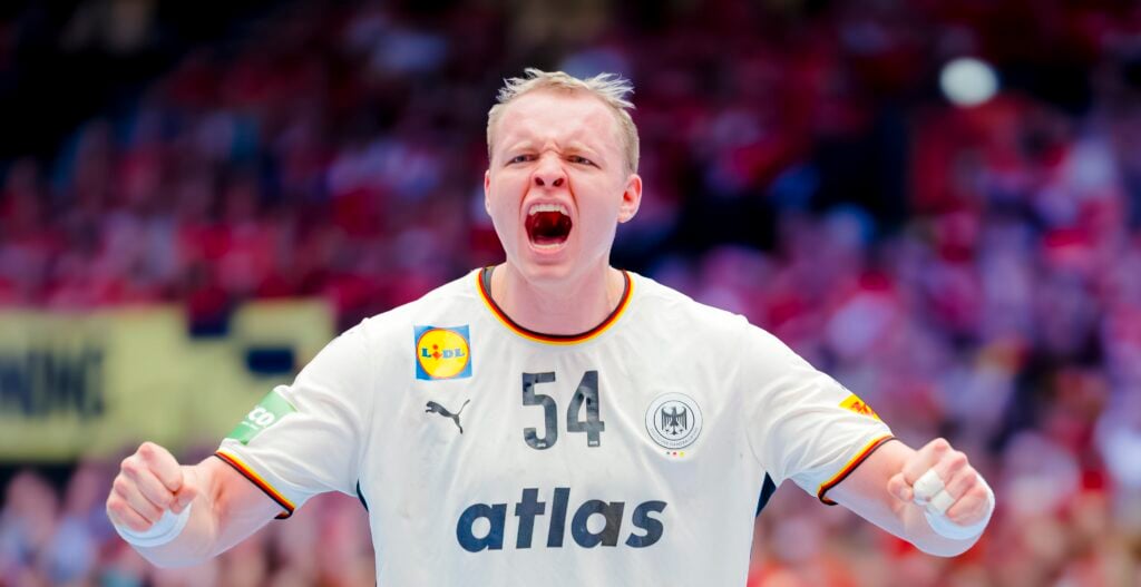 Ein Handballspieler, der ein weißes Trikot mit der Nummer 54 und dem Emblem Deutschlands trägt, schreit energisch auf dem Spielfeld, während im Hintergrund verschwommene Zuschauer in Rot zu sehen sind.