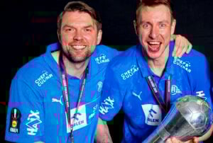 Zwei Männer in passenden blauen Sporthemden und Medaillen lächeln freudig, der eine hat den Arm um die Schulter des anderen gelegt und hält eine silberne Trophäe vor einem schwarzen Hintergrund.