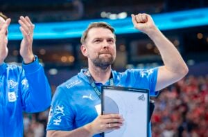 Ein Mann in einem blauen Sporthemd hebt triumphierend die rechte Faust, während er ein Klemmbrett hält. Er steht in einer Arena und trägt eine Medaille, während im Hintergrund eine Menschenmenge zu sehen ist.