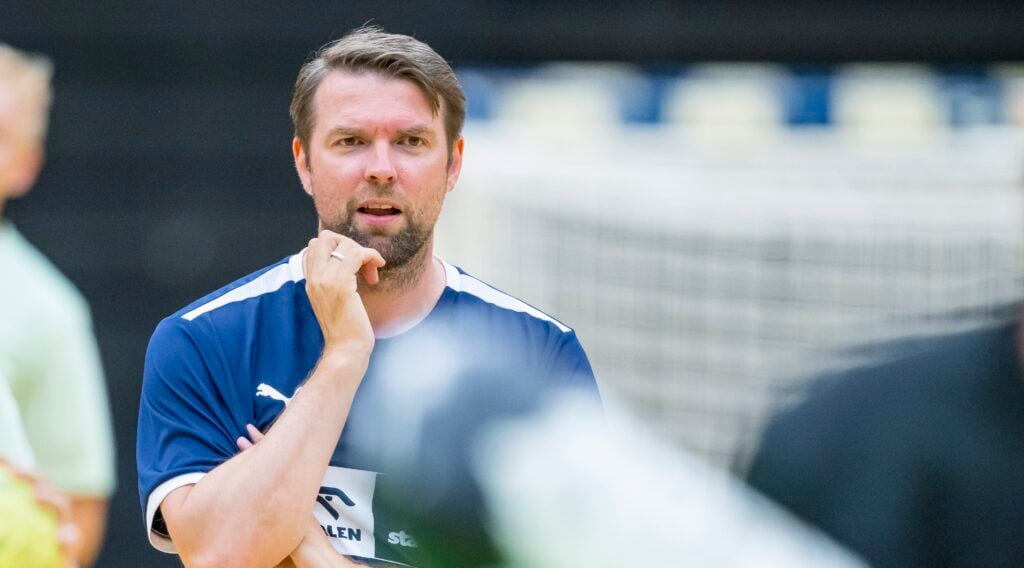 Ein Mann in einem blauen Sporthemd steht mit der Hand am Kinn und schaut nachdenklich. Der Hintergrund ist unscharf und zeigt ein Tornetz und eine weitere unscharfe Person.