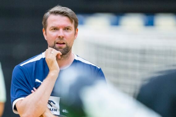 Ein Mann in einem blauen Sporthemd steht mit der Hand am Kinn und schaut nachdenklich. Der Hintergrund ist unscharf und zeigt ein Tornetz und eine weitere unscharfe Person.