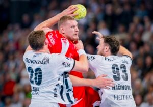 Ein Handballspieler in einem roten Trikot springt, um den Ball zu werfen, während zwei Verteidiger in weißen Trikots versuchen, ihn während eines rasanten Spiels zu blockieren. Im Hintergrund ist eine unscharfe Menschenmenge zu sehen.