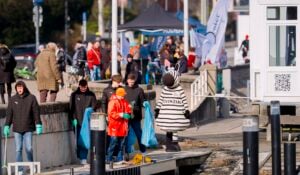 Eine Gruppe von Menschen, darunter eine Person in einem Zebrakostüm, säubert das Ufer von Müll. Mehrere Personen halten blaue Müllsäcke, und einige tragen Handschuhe und helle Jacken. Im Hintergrund sind Veranstaltungszelte und Fahnen zu sehen.
