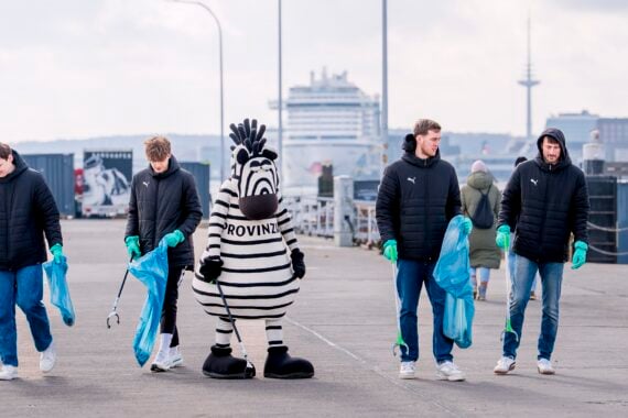 Vier Personen in schwarzen Jacken und grünen Handschuhen sammeln mit Greifern und blauen Säcken Müll auf einem Pier ein, begleitet von einer Person in einem schwarz-weiß gestreiften Zebra-Maskottchenkostüm. Im Hintergrund ist ein Kreuzfahrtschiff zu sehen.
