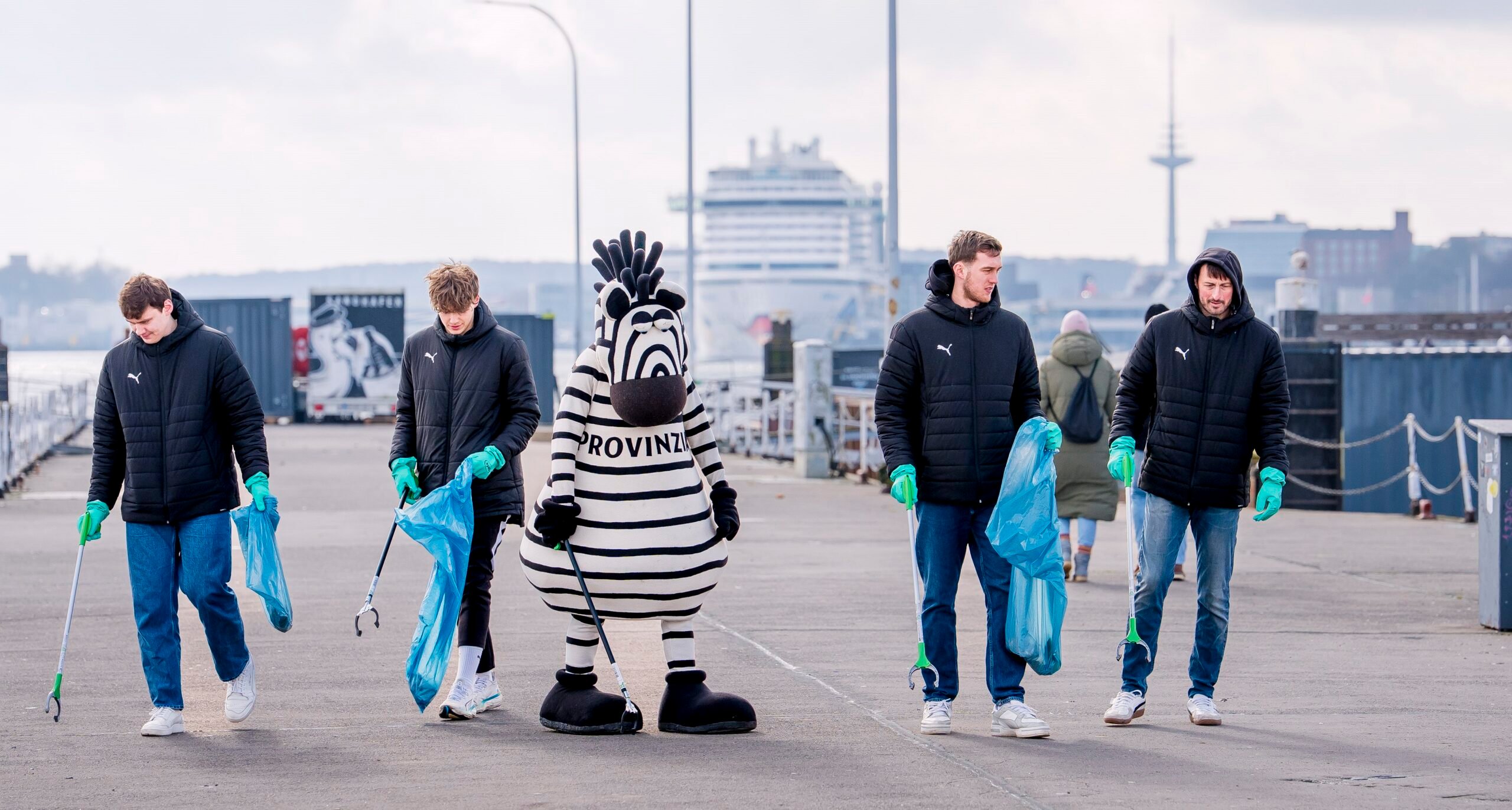 Vier Personen in schwarzen Jacken und grünen Handschuhen sammeln mit Greifern und blauen Säcken Müll auf einem Pier ein, begleitet von einer Person in einem schwarz-weiß gestreiften Zebra-Maskottchenkostüm. Im Hintergrund ist ein Kreuzfahrtschiff zu sehen.