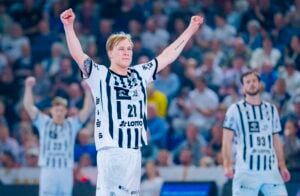 Ein Handballspieler in einem schwarz-weiß gestreiften Trikot reißt auf dem Spielfeld jubelnd beide Arme in die Höhe, während zwei Mannschaftskameraden in ähnlichen Trikots im Hintergrund ebenfalls die Arme in die Höhe reißen. Das Publikum ist unscharf.