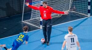 Ein Handballtorwart in einem roten Trikot steht mit ausgestreckten Armen und schützt das Tor, während ein Spieler in Blau mit der Nummer 21 nach vorne fällt und ein Spieler in Weiß mit der Nummer 11 in der Nähe auf einem blauen Feld steht.