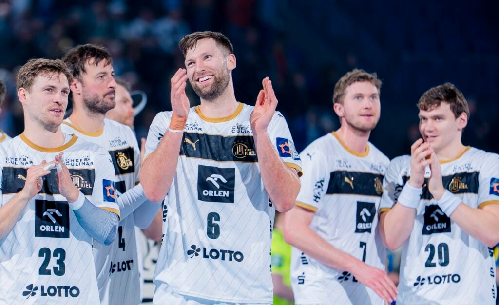 HANDBALL 2025/26: THW Kiel - Bidasoa Irun Fünf männliche Handballspieler in weißen und schwarzen ORLEN-Trikots stehen zusammen auf dem Spielfeld, lächeln und klatschen und wirken nach einem Spiel glücklich und feierlich. Der Hintergrund ist mit Zuschauern unscharf.