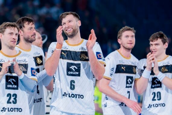 Fünf männliche Handballspieler in weißen und schwarzen ORLEN-Trikots stehen zusammen auf dem Spielfeld, lächeln und klatschen und wirken nach einem Spiel glücklich und feierlich. Der Hintergrund ist mit Zuschauern unscharf.