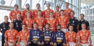 Eine Herren-Handballmannschaft posiert in der Halle in orangefarbenen Trikots mit Sponsorenlogos. Sie sind in drei Reihen mit Trainern und Betreuern, einige in marineblauen Hemden, aufgestellt, die zusammen stehen und sitzen und in die Kamera schauen.