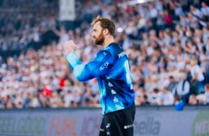 Ein männlicher Handballspieler in einem blauen Trikot steht mitten im Jubel mit erhobener Faust auf einem Spielfeld vor einer verschwommenen, jubelnden Menge.