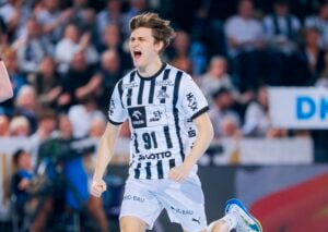 Ein männlicher Handballspieler in einem schwarz-weiß gestreiften Trikot mit der Nummer 91 jubelt auf dem Spielfeld mit offenem Mund und geballter Faust, während die Zuschauer im Hintergrund zuschauen.