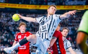 Ein männlicher Handballspieler in einem schwarz-weißen Trikot springt, um den Ball zu werfen, während seine Gegner in roten Trikots ihn verteidigen. Das Geschehen spielt sich in einer überfüllten Halle ab.
