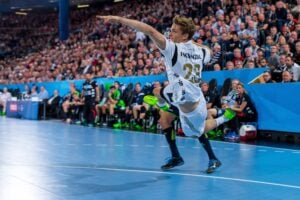 Ein Handballspieler in einem weißen Trikot springt mit ausgestrecktem Arm in die Luft, um den Ball zu werfen, während die Zuschauer in der vollbesetzten Arena das Spiel aufmerksam verfolgen.