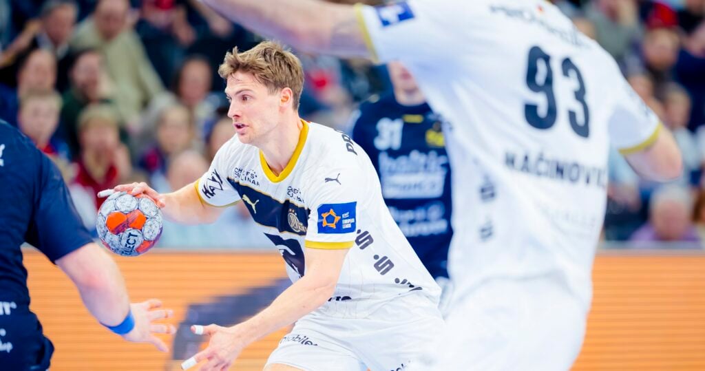 Ein Handballspieler in einem weißen Trikot bereitet sich darauf vor, den Ball zu werfen, während er von Spielern in dunklen Trikots während eines intensiven Spiels verteidigt wird, während die Zuschauer im Hintergrund zuschauen.