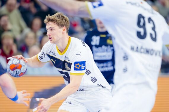 Ein Handballspieler in einem weißen Trikot bereitet sich darauf vor, den Ball zu werfen, während er von Spielern in dunklen Trikots während eines intensiven Spiels verteidigt wird, während die Zuschauer im Hintergrund zuschauen.