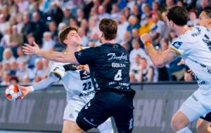Ein Handballspieler in einem weißen Trikot versucht, den Ball zu passen, während er von einem Verteidiger in einem dunklen Trikot während eines rasanten Spiels geblockt wird, während die Zuschauer im Hintergrund zuschauen.